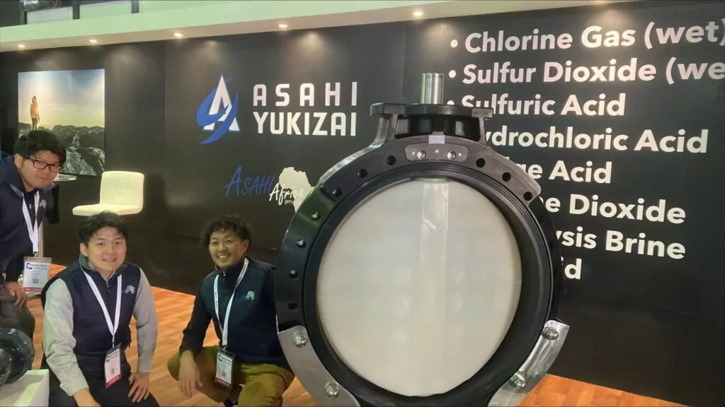 Employees of Asahi Africa kneeling down next to a large 900mm thermoplastic diaphragm valve, with a black wall and text and the Asahi Africa logo.